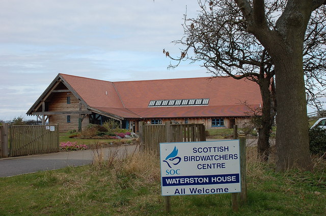 Wooden building with a reddish roof beyond a short driveway and trees. The sign in the front reads "Scottish Birdwatchers Centre - Waterson House - All Welcome."