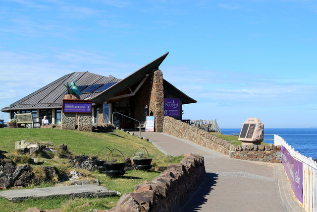Stone building with an angular roof up a driveway and set right on the coast; blue skies and water in the background.