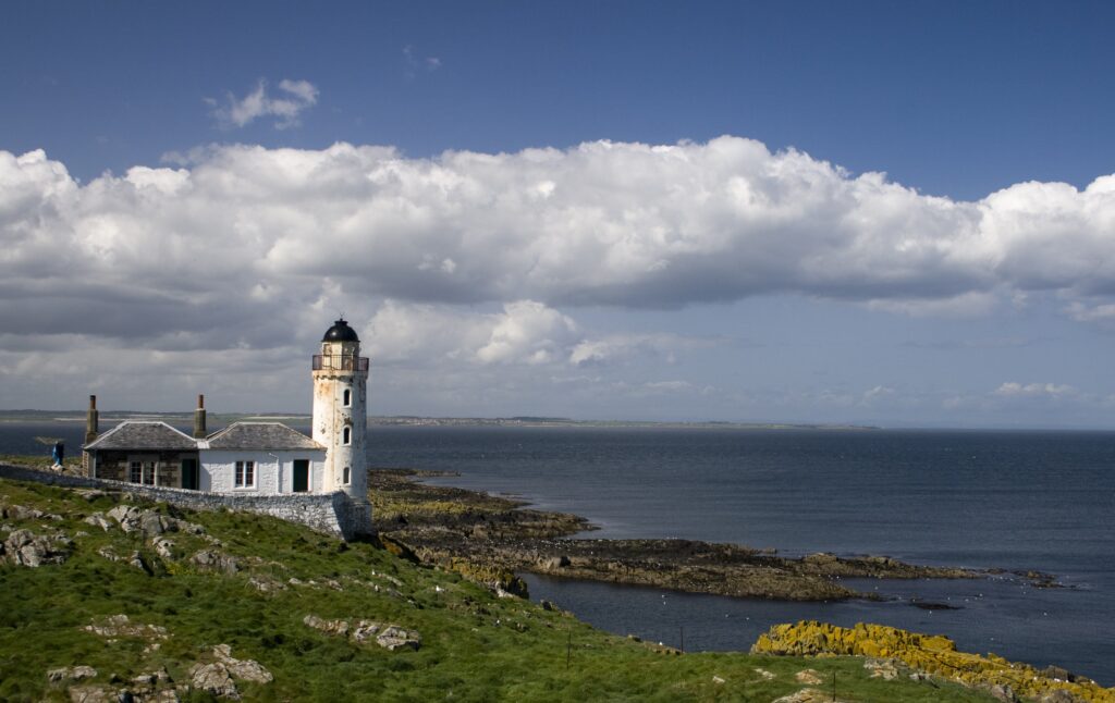 White lighthouse on a rocky coastline with greens in the foreground and blue skies and sea in the background.