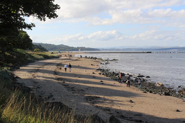 Shoreline with green foliage on the left, multiple people walking on tan, rocky sand, and a protruding dock going far into the water.