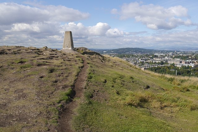 Green hill with a path to a tall cement structure under a cloudy blue sky, with city views in the background.