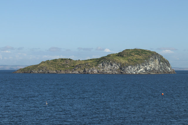 Rocky island with green foliage; dark blue waters in the foreground and light blue skies in the background.