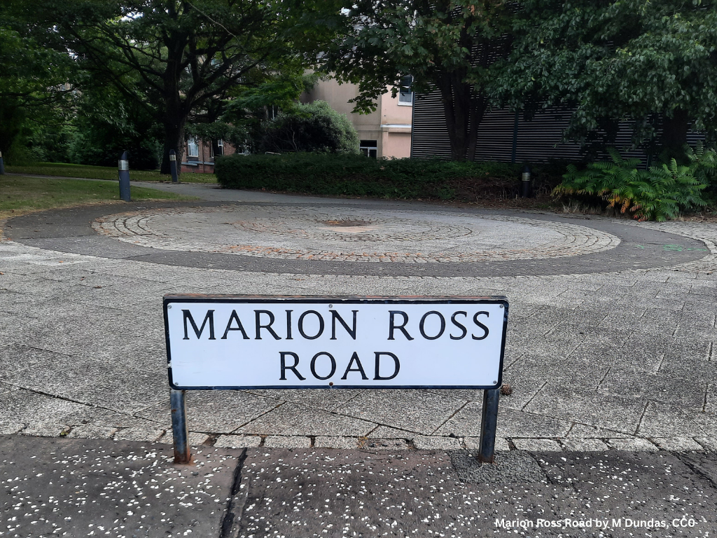 A short white street sign on two legs that says Marion Ross Road.  The sign is in front of a paved brick circle and trees.  