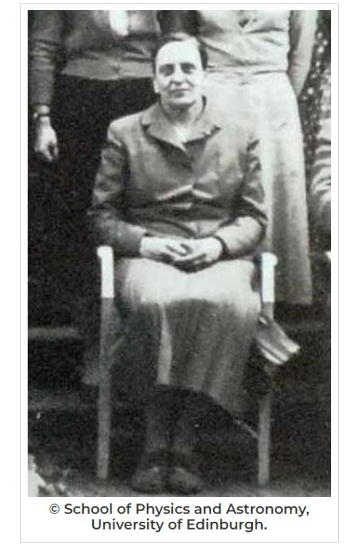 A black and white photo (circa 1940s-1950s) of a dark-haired, strong-featured woman,  wearing a suit and skirt and sitting in a chair as part of a group photo.