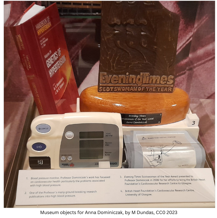 A display of medical equipment and books, including a blood pressure monitor, a book on the genetics of hypertension, and a Scotswoman of the year award. 