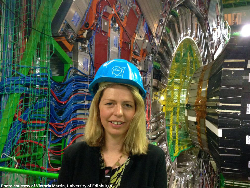 A light-haired woman wearing a dark suit and blue hard hat with a CERN logo in front of a background of colourful cables, wires and equipment.