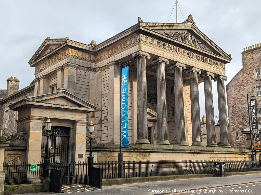 A tall light stone building with six columns, a decorated pediment, and a blue Welcome banner out front. There is a small plaque at the gate on the left, behind two lamp posts.