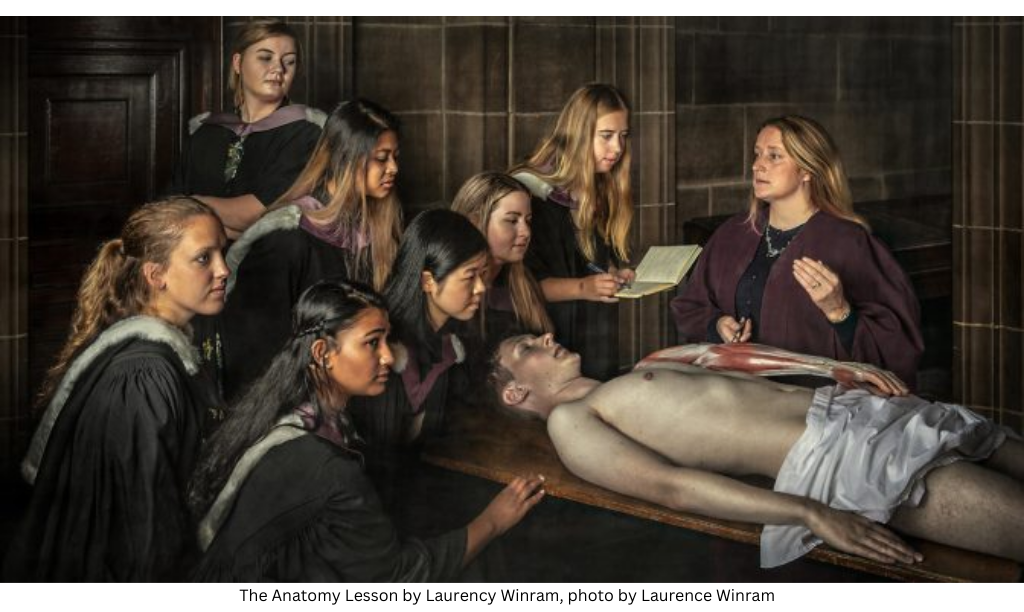 A group of seven young women in black graduation gowns around a table with a cadaver on it and a female instructor giving an anatomy lesson the arm.