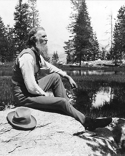 Picture of John Muir, an older man with a long beard, sitting on a rock at a lake surrounded by pinte trees, staring into the distance with his hat next to him on the rock.