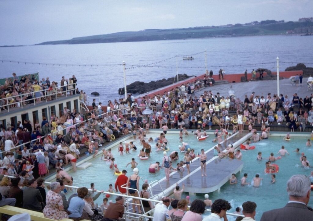 Modern photo of pool with people swimming, coastline in the background.