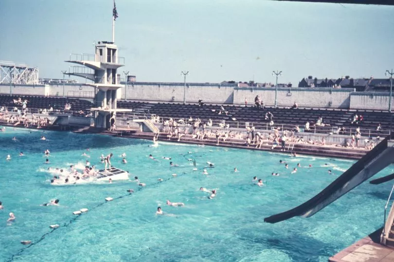 Open air pool with people swimming; high dive platforms and bleachers in the background.