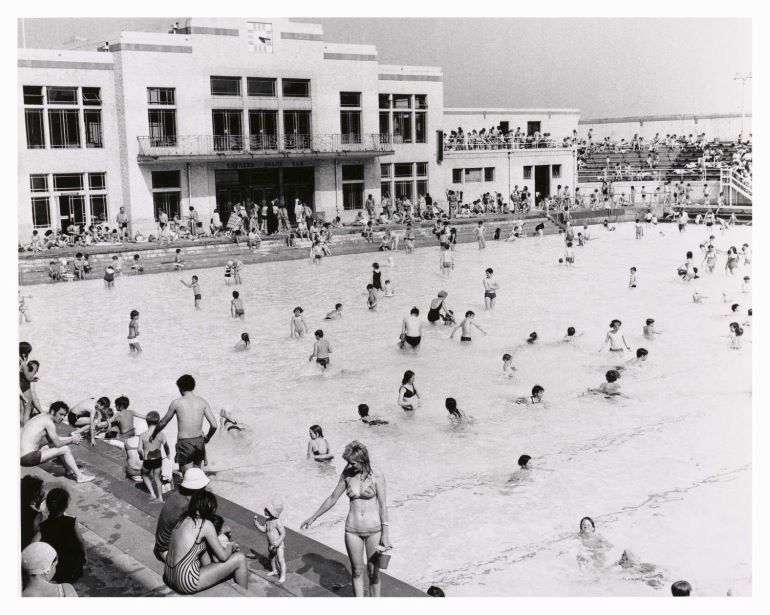 Black and white photo of pool with people swimming; white buildings in the background.