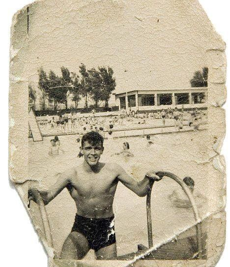 Sepia photo of young man climbing out of open air pool using the ladder; people swimming, buildings, and trees in the background.