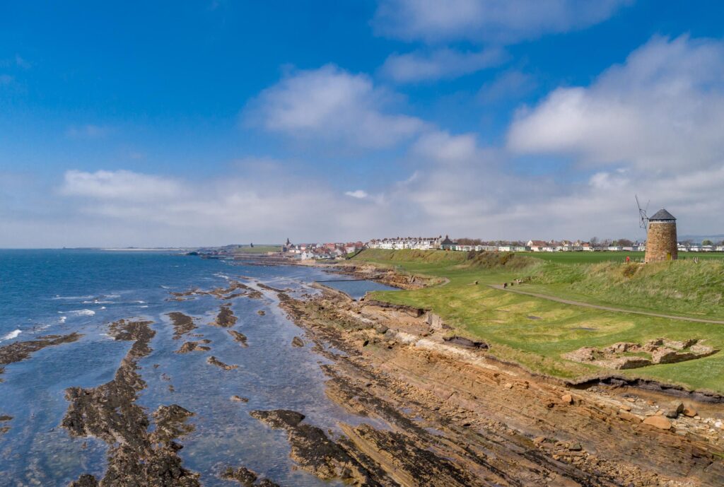 Rocky coastline with green grass and a stone windmill on the right side, and blue waters on the left; brilliant blue sky with clouds in the background.