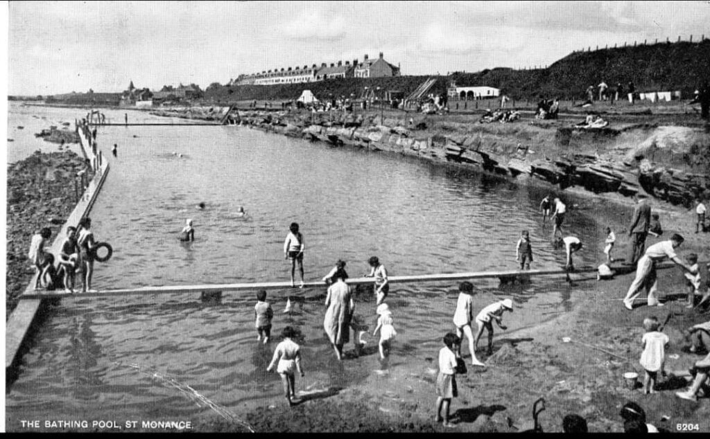 Black and white photo of pool with people swimming; rocky coastline and row buildings in the background.