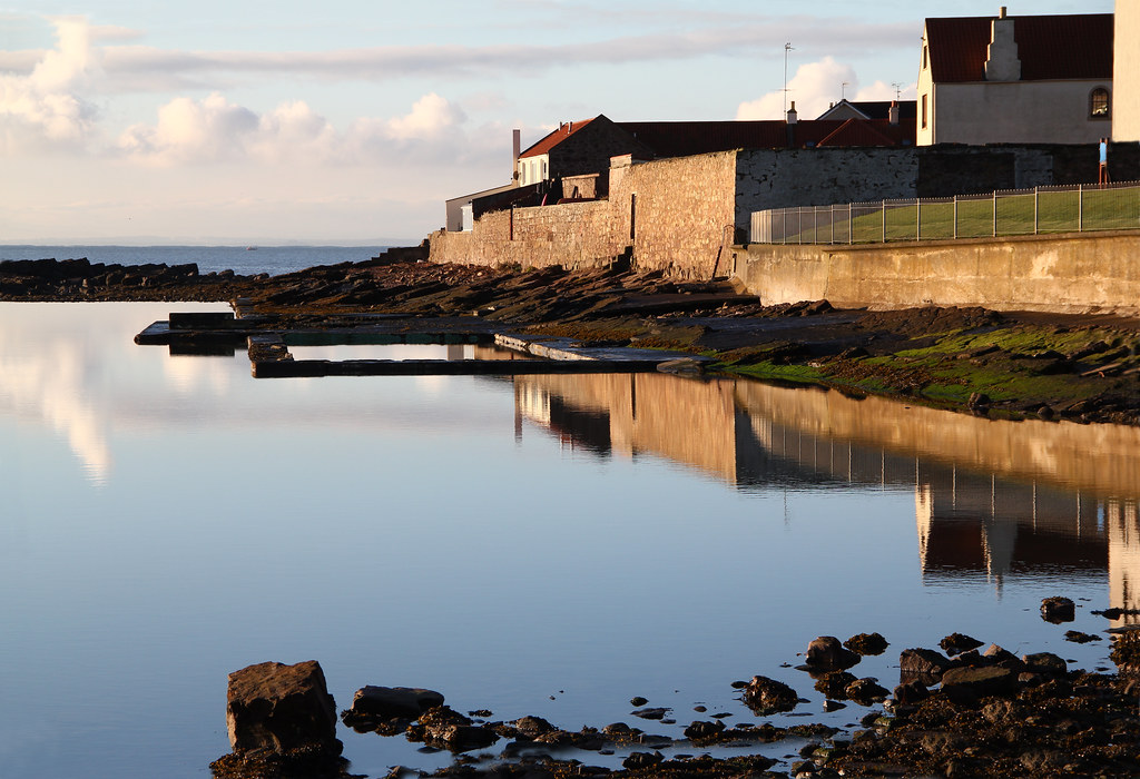 Still, reflective tidal pool with rocky shore and buildings in the background.