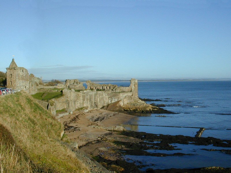 Tidal pool set against the back of a stone castle and wall with a green, grassy sloping hill; blue ocean in the foreground and background.
