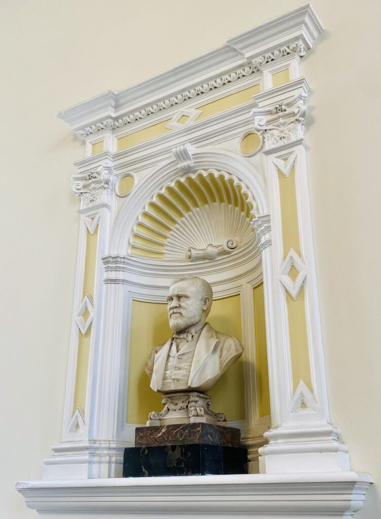 Stone carving bust of a man with a beard, wearing a suit, inside an ornate yellow and white wall inset.
