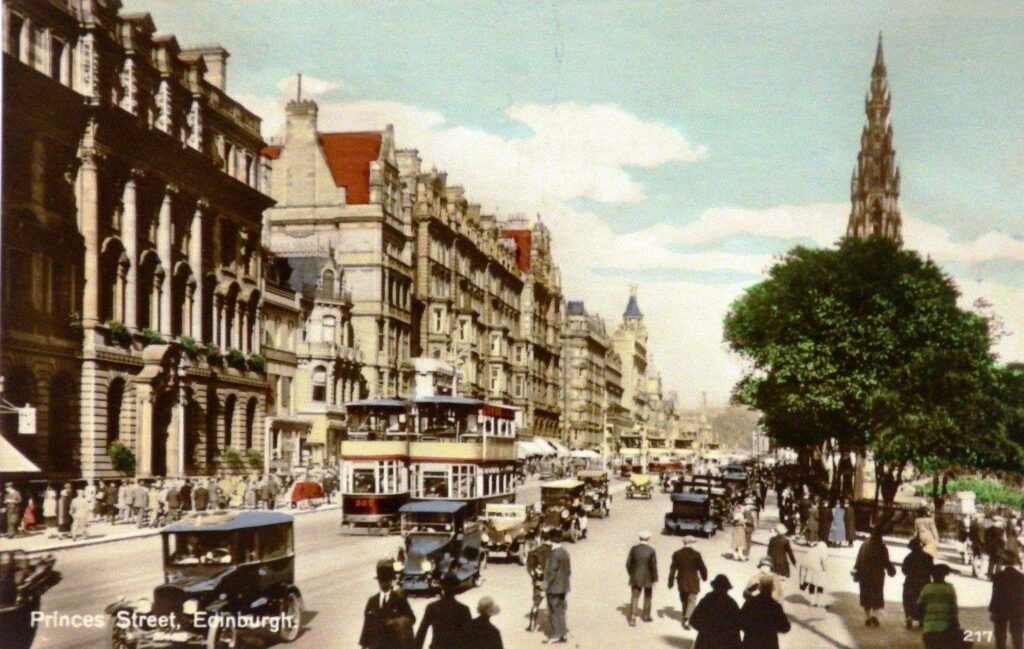 Busy street view of Princes Street with early model cars and buses, pedestrians, buildings, trees, and the Scott Monument in the background.