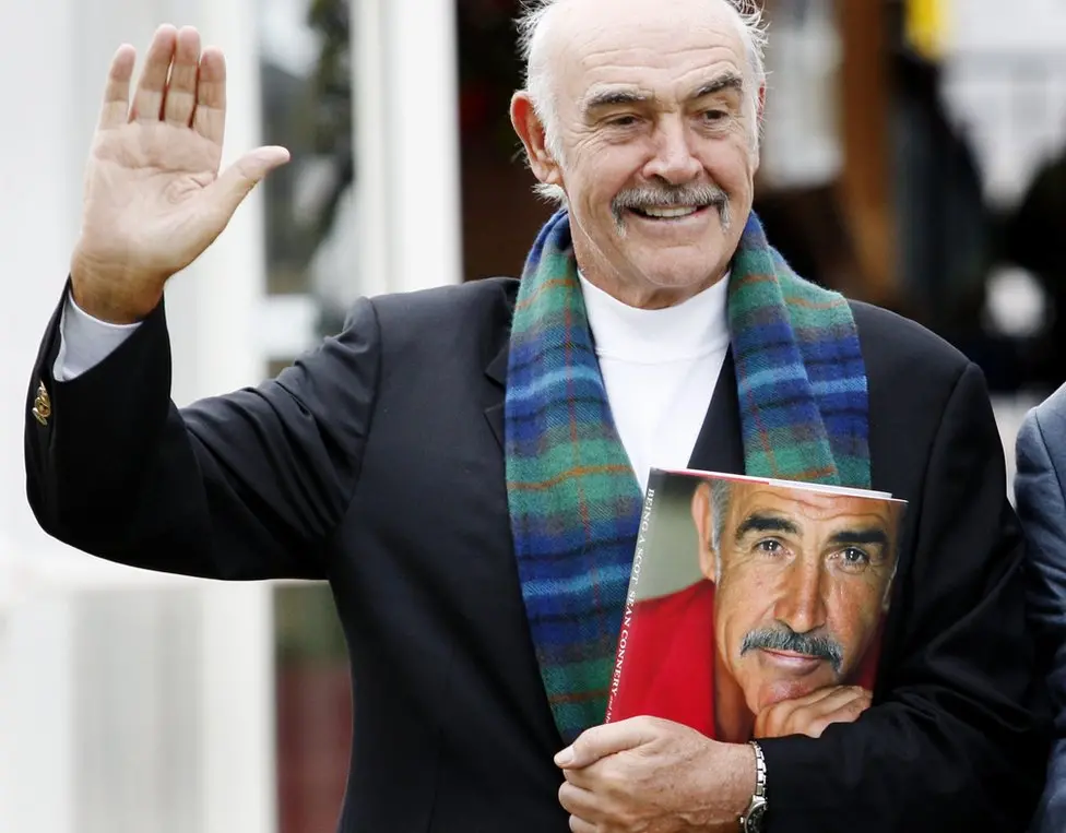 Older balding gentleman with a mustache waving, wearing a navy blazer, white mock turtleneck, blue-green tartan scarf, and carrying a book with his younger face on it.