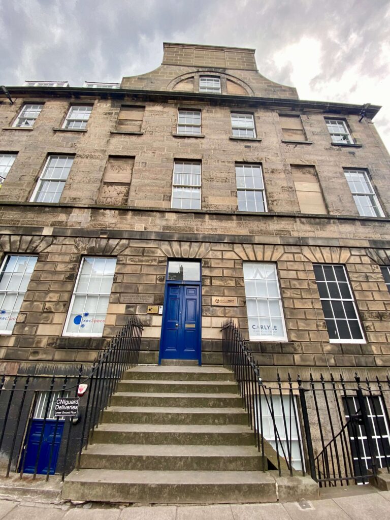 Tall limestone building with multiple windows, unique roofline, and royal blue door atop a short staircase.
