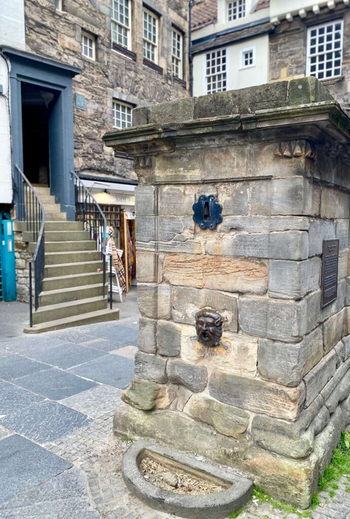 On the right: large, squared stone cistern with a metal open-mouthed face toward the bottom; on the left: curved staircase in the background.