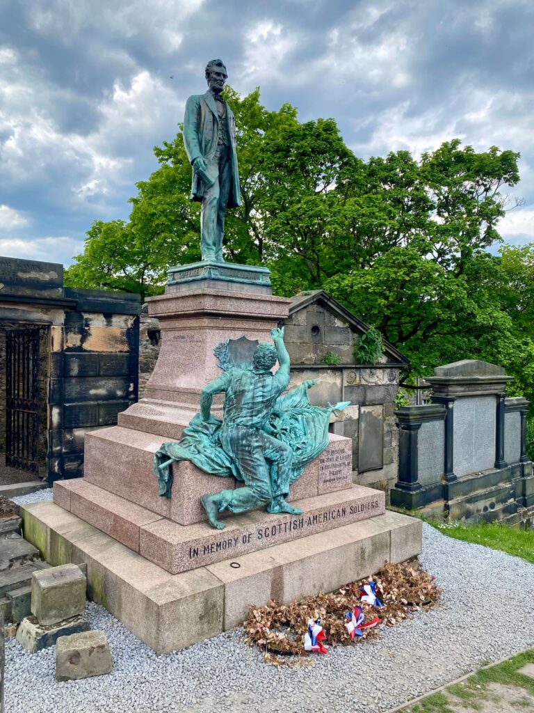 Granite and bronze (teal) statue with Abraham Lincoln atop and a slave reaching upward, situated in a cemetery setting surrounded by gravel and dried wreaths.