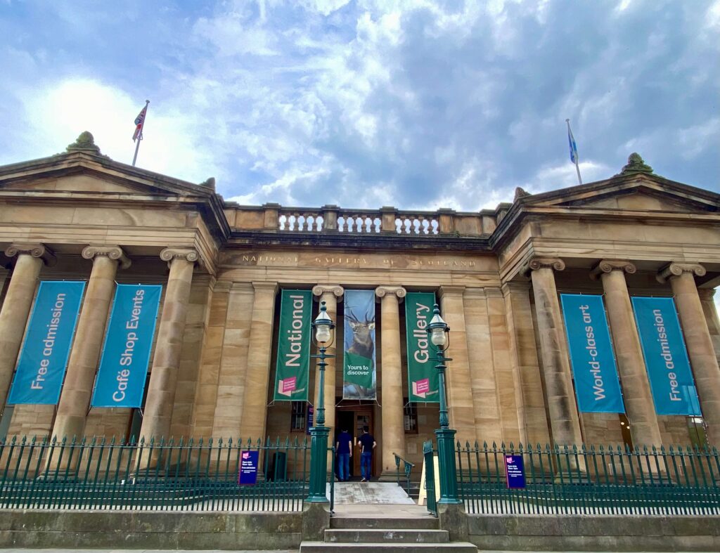 Limestone building with blue and green banners, green fence, light posts, flags and steps up.