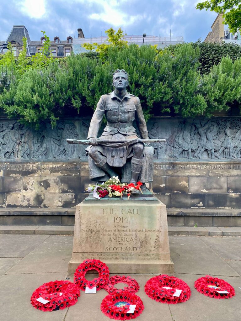 Bronze statue of a seated soldier in uniform with a gun; frieze and foliage behind him; red poppy wreaths in front.