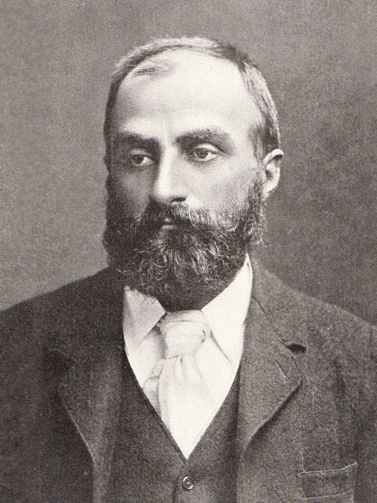 Black and white portrait of a beaded and early balding man wearing a dark jacket and light shirt and tie. 