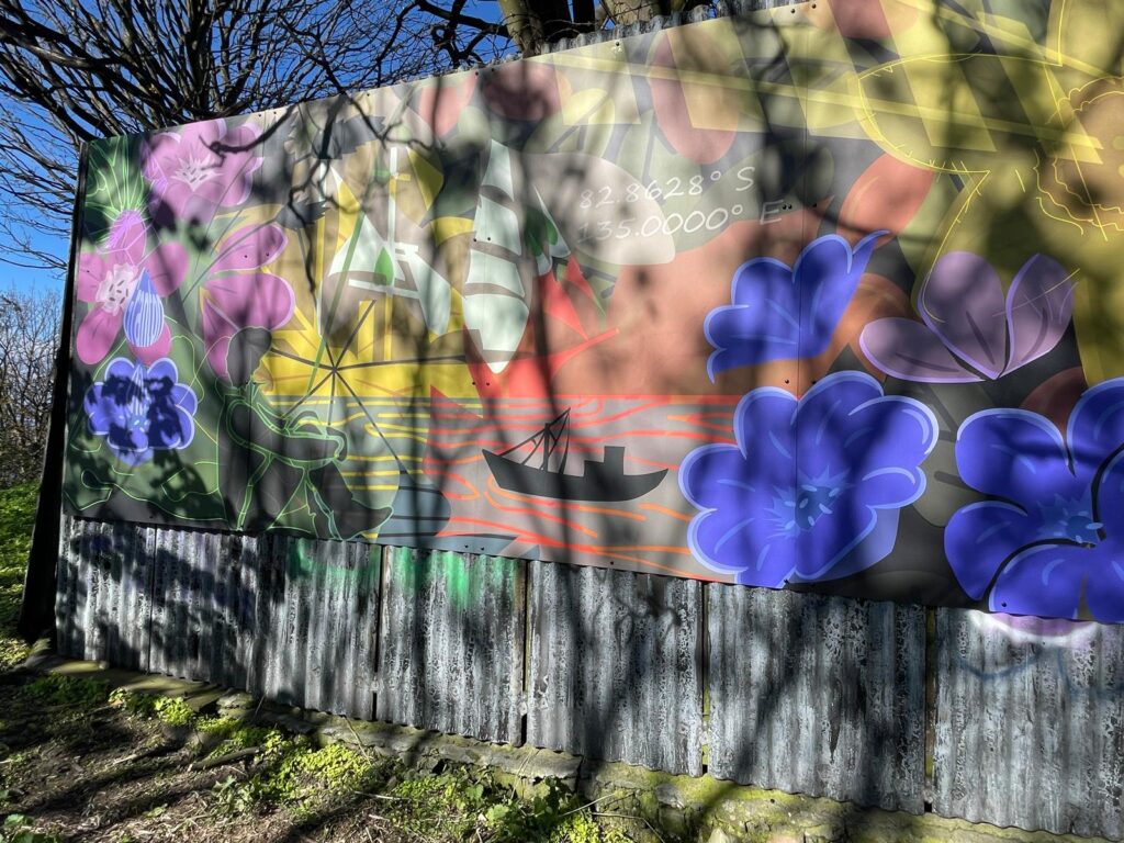 Colorful horizontal mural with purple and yellow flowers, a black boat, and seated person in front of trees with no leaves and under a blue sky.